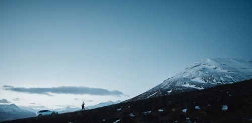 Wide shot of a person walking up a hillside near a white SUV, set against a backdrop of mountains and a clear sky.
