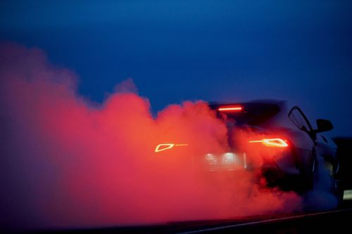 Grey sports car drifting at night with red smoke billowing from the tires, tail lights and headlights reflecting in the dark.