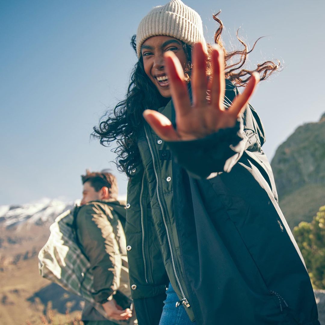 Smiling woman in winter hat and coat reaching out, with a man and snowy mountains in the background on a sunny day.
