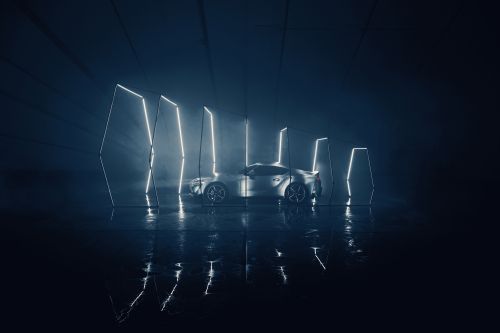 Silver sportscar in a dark garage with neon light tubes reflecting on wet floor, under a shadowy light. Automotive promotional image.