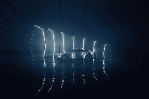 Silver sportscar in a dark garage with neon light tubes reflecting on wet floor, under a shadowy light. Automotive promotional image.