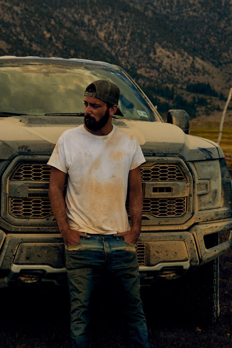 Man in dirty t-shirt leans against a muddy pickup truck with mountains in the background. He wears a baseball cap and blue jeans.
