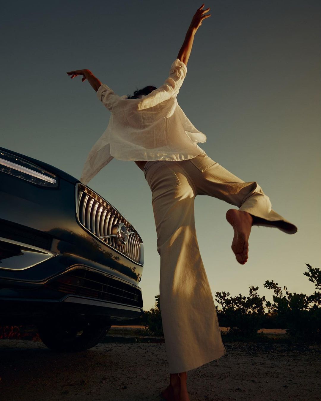 Woman in casual clothing balances barefoot next to a dark blue Volvo SUV with the sun low in the sky at daytime.