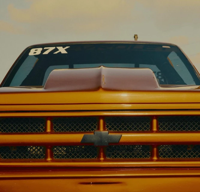 Front view of an orange pickup truck, showing the grille, headlights, hood, and windshield with a sky background. Racing number on glass.