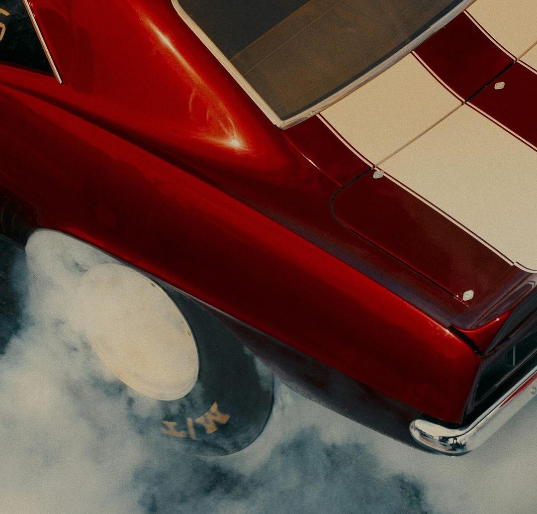 Overhead shot of a red vintage Chevrolet Camaro with a white stripe and smoke rising from the tires after a burnout.