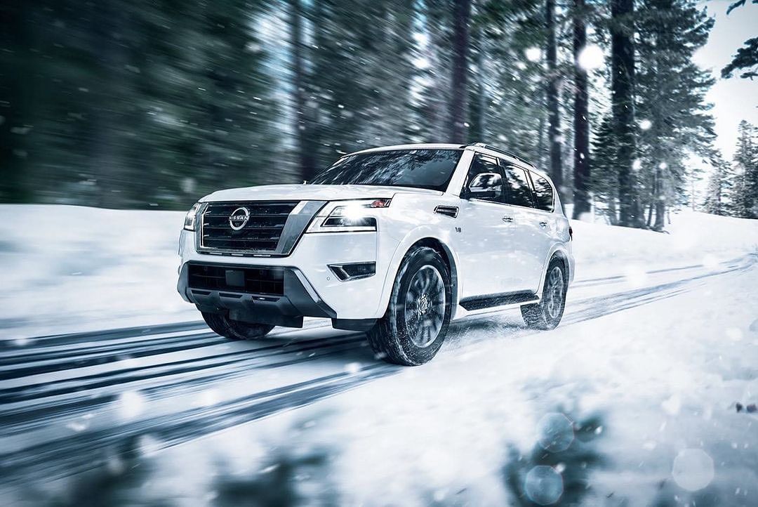 White SUV driving on snow covered road, surrounded by winter trees. Vehicle is in motion with snow and ice visible on the ground.