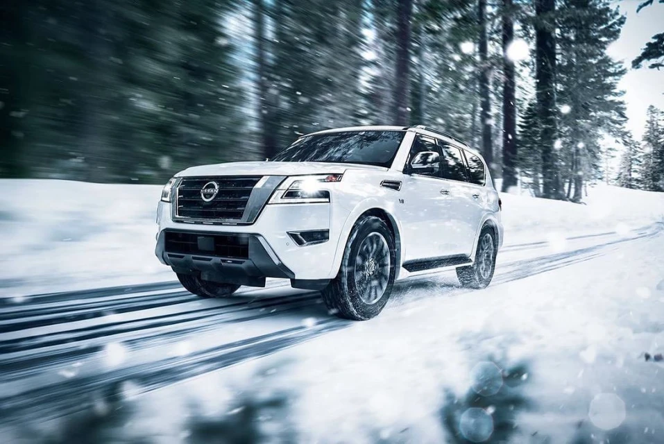 White SUV driving on snow covered road, surrounded by winter trees. Vehicle is in motion with snow and ice visible on the ground.