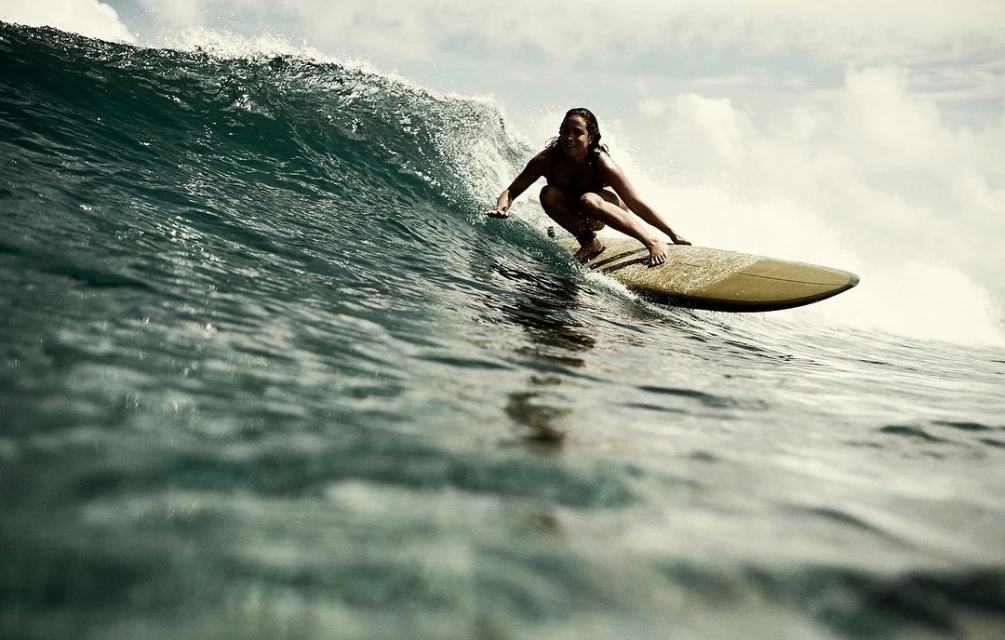 Woman in swimsuit riding a surfboard on a large ocean wave with cloudy sky, capturing the essence of water sports and summer adventure.