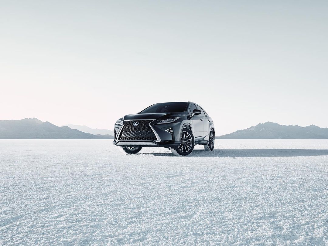 Dark grey Lexus SUV parked on a flat white surface with mountains in the distance under a bright sky, automotive product shot.