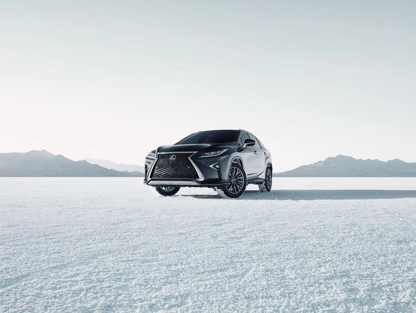 Dark grey Lexus SUV parked on a flat white surface with mountains in the distance under a bright sky, automotive product shot.