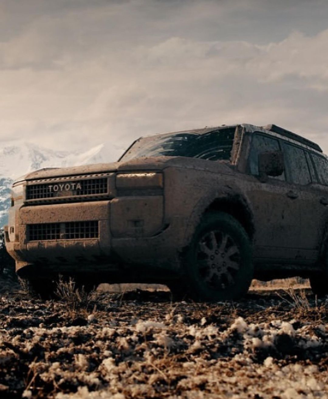 Off-road mud-covered Toyota SUV in a rugged terrain landscape with snowy mountains in the distance under an overcast sky.