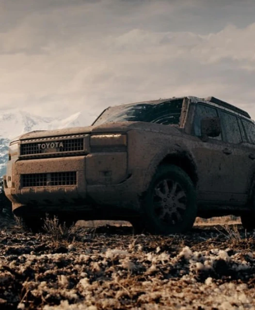Off-road mud-covered Toyota SUV in a rugged terrain landscape with snowy mountains in the distance under an overcast sky.