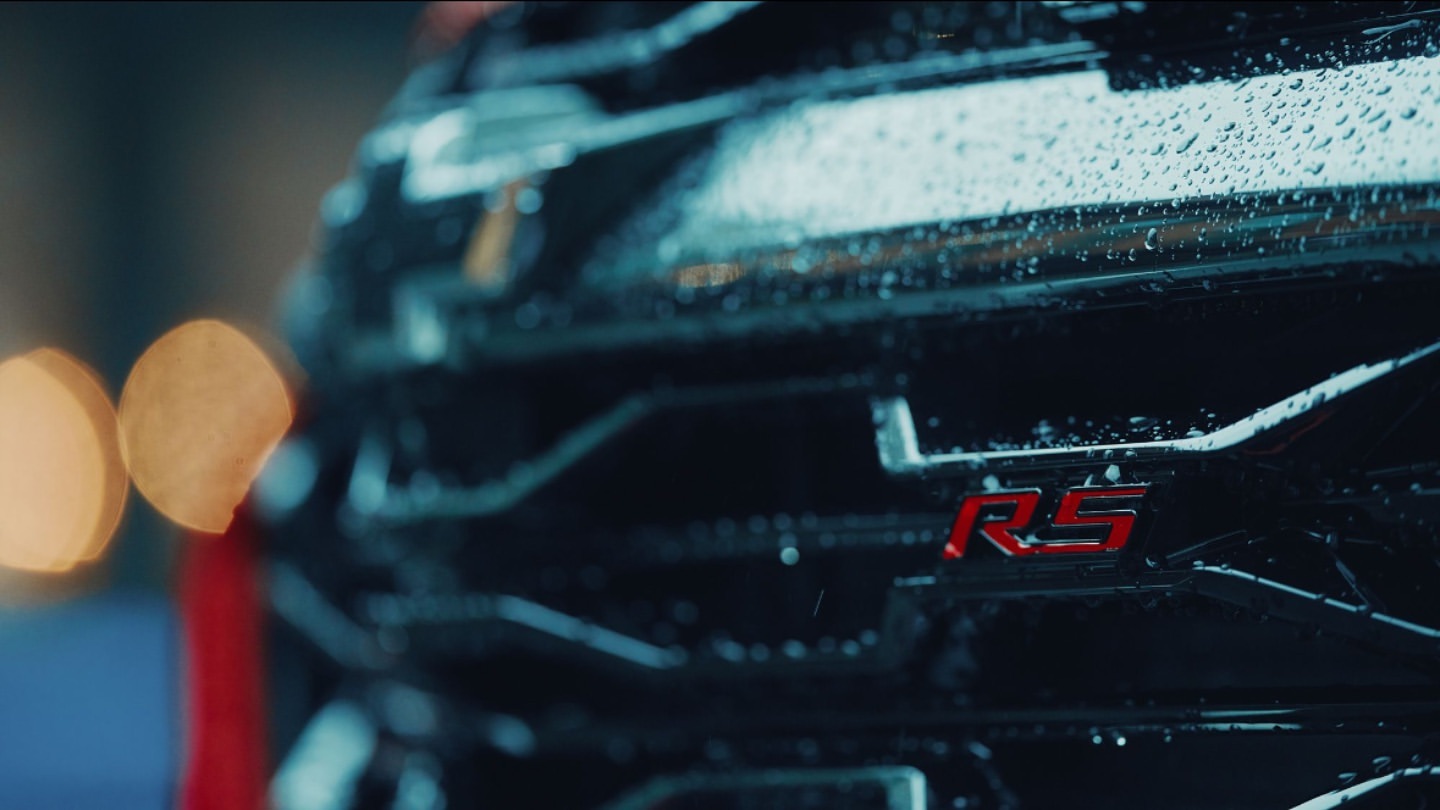 Close-up of a black car's grill with water droplets and a red 'RS' emblem, creating a moody and detailed automotive shot.