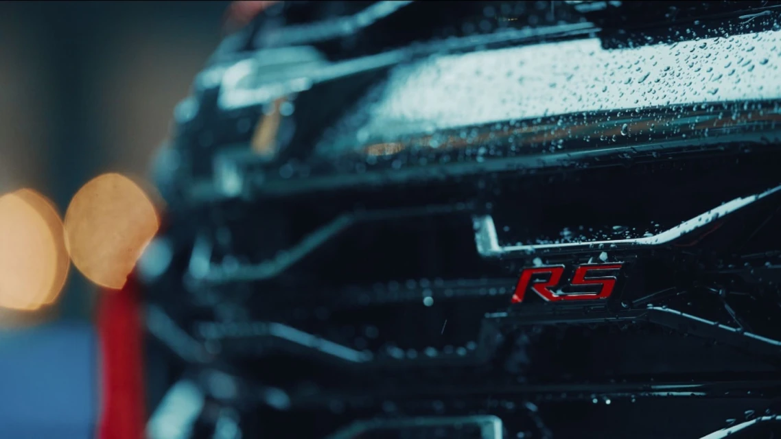 Close-up of a black car's grill with water droplets and a red 'RS' emblem, creating a moody and detailed automotive shot.