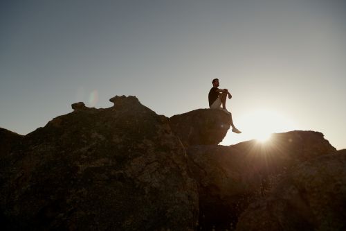 A man sits atop rocks against the sunlight. The figure is silhouetted and contemplative amidst a clear sky and rocky terrain.
