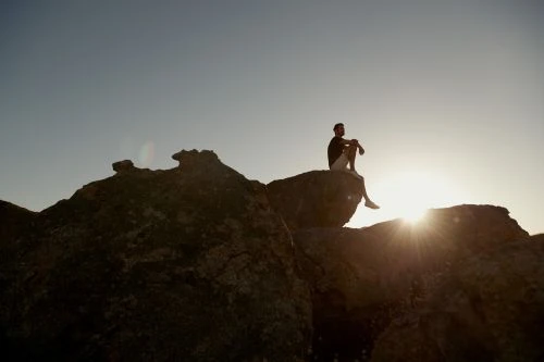 A man sits atop rocks against the sunlight. The figure is silhouetted and contemplative amidst a clear sky and rocky terrain.