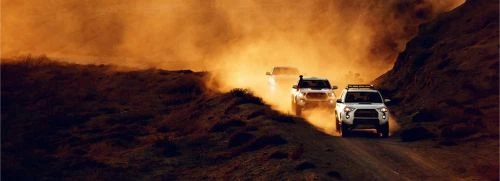 Three white SUVs driving on a dusty desert road between two dark, rocky hills kicking up dust in the diffused sunlight.