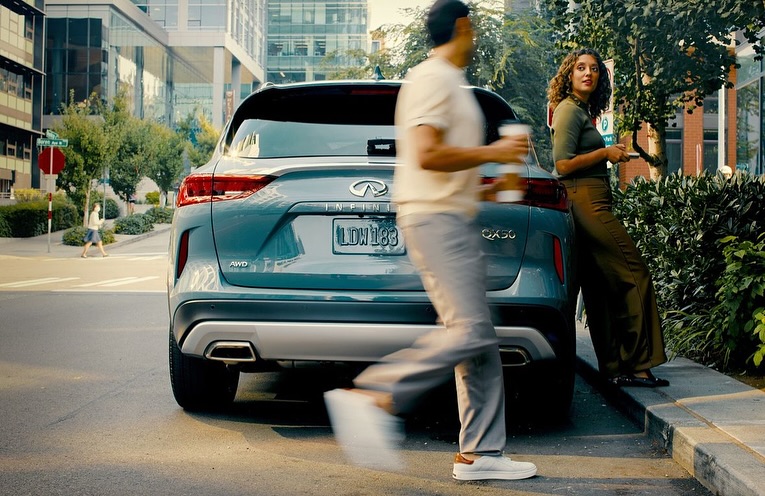 Turquoise Infiniti QX50 SUV parked on a city street with a man walking by holding a coffee and a woman standing next to it.