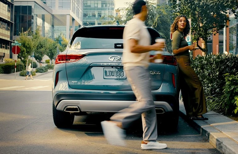 Turquoise Infiniti QX50 SUV parked on a city street with a man walking by holding a coffee and a woman standing next to it.