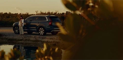 Woman standing near a dark blue Volvo XC90 on a sandy shore near a body of water at sunset; lush green vegetation surrounds the car.