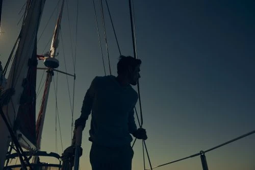 Silhouette of a man stands on a sailboat under a dusky sky. The boat's mast and sails are partially visible, with a serene, nautical ambiance.