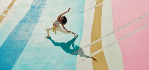 A woman in athletic wear does a dynamic pose on a colourful painted surface. Viewed from overhead, her shadow is visible on the ground.