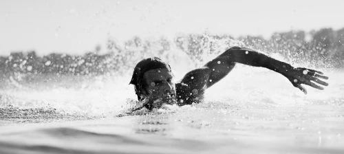 Monochrome shot of a swimmer in water, arm raised, creating splash against bright sky, displaying energy and athletic skill.