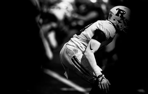 High-contrast monochrome photograph of an American football player in uniform wearing a helmet crouched on a field.