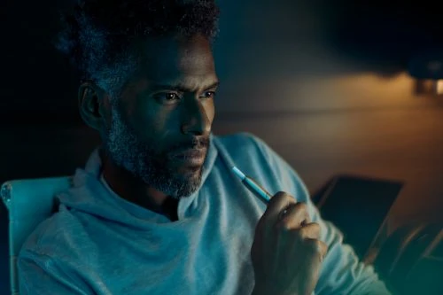 Man in hoodie holds vape pen in indoor setting with blurred background, wearing casual attire, looking off-camera with thoughtful expression.