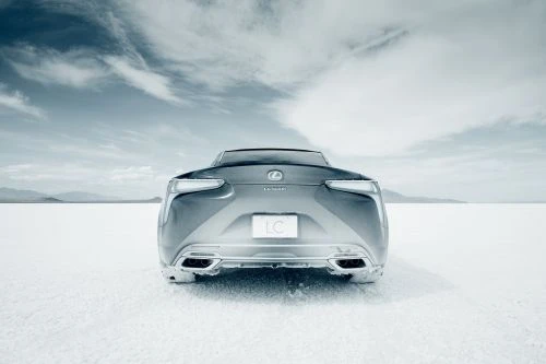 Rear view of a grey Lexus LC coupe car on a white surface under a cloudy sky, showcasing the car's design and outdoor setting.