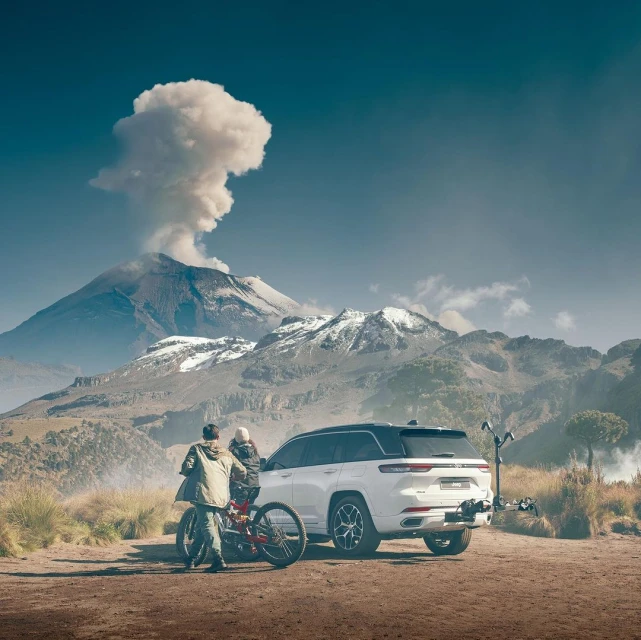 A white SUV with a bike rack and two people stands before a dormant, snow-capped volcano in a mountainous, grassy landscape.
