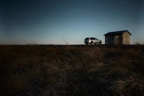 Silver SUV parks near a small, rustic wooden structure in a field of brown grass against a blue sky. Automotive outdoor shot.