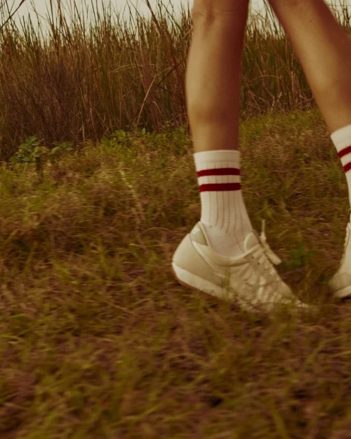 A close-up shot of a person wearing white sneakers and striped socks walking through a grassy field on a sunny day outdoors.
