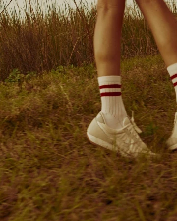 A close-up shot of a person wearing white sneakers and striped socks walking through a grassy field on a sunny day outdoors.