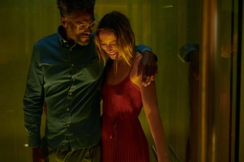 A man and woman embrace affectionately indoors, with the woman in a red dress and the man in a blue shirt, bathed in atmospheric lighting.