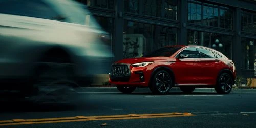 A red Infiniti QX55 coupe SUV driving on a city street with blurred motion, showcasing automotive design and urban transport.