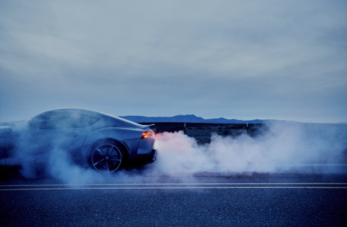 Gray sports car drifting on a dark asphalt road surrounded by a cloud of white smoke against a backdrop of cloudy sky and distant mountains.