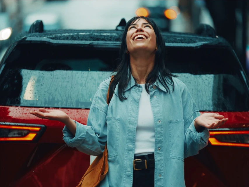 A smiling woman in a denim jacket stands in the rain with a red SUV behind her in an urban setting on an overcast day.