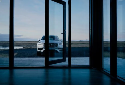 Silver SUV parked outside a modern house with an open glass door, reflecting the landscape and sky in a serene, minimalist setting.