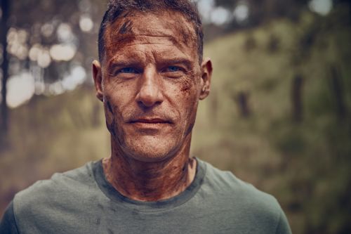 Close-up shot of a muscular adult male with dirt on his face and head, wearing a green shirt and standing in an outdoor setting with trees.
