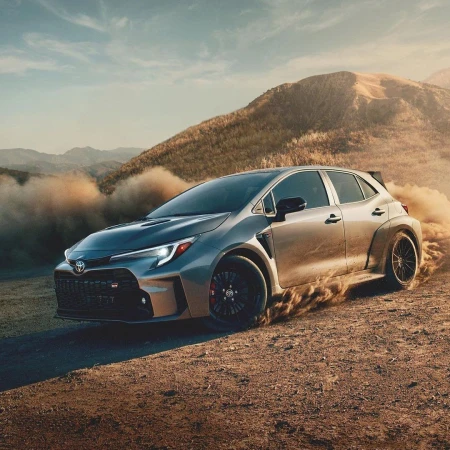 Silver Toyota GR Corolla hatchback speeds across a dirt road in a mountainous landscape, kicking up dust. Automotive product shot.