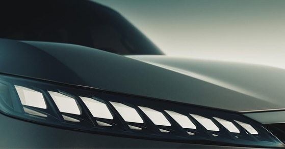 Close-up of the front of a gray car showcasing a modern headlight design with geometric shapes against a soft, neutral backdrop.