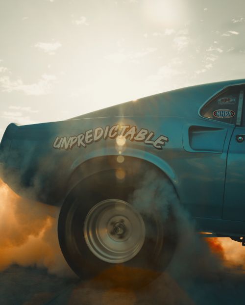 The rear of a light blue classic Ford Mustang drag car does a burnout, tires smoking against a sun-drenched sky at daytime.