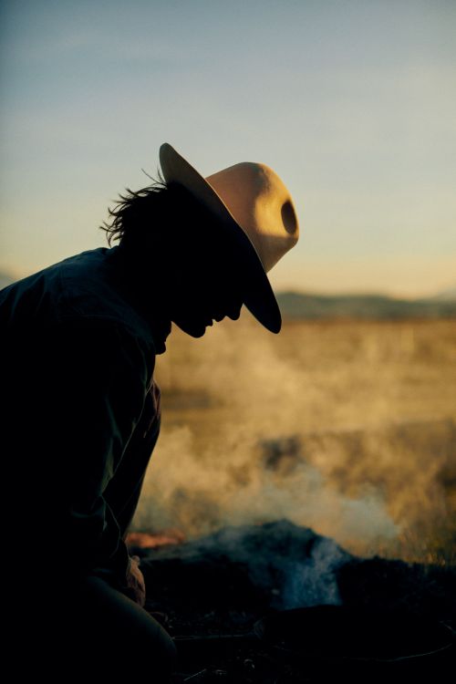 Silhouette of a person in a cowboy hat sitting near a campfire with smoke, against a dusky sky and expansive grassy landscape.