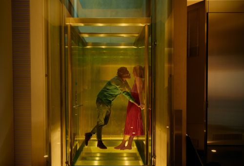 A man and woman are embracing in a transparent elevator, visible through glass walls with warm, soft lighting creating a romantic atmosphere.