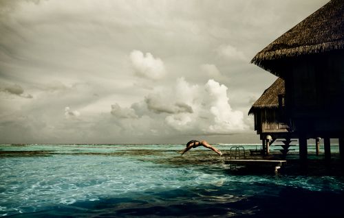Person dives into turquoise ocean water near a thatched-roof bungalow with cloudy skies overhead. Summer tropical travel destination.