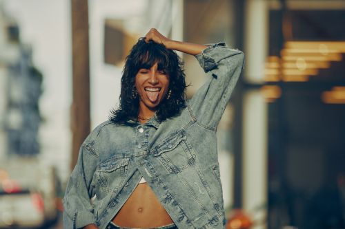 A young adult female playfully sticks out her tongue while posing outdoors, wearing a denim jacket and showcasing her midriff in a light-filled, urban setting.