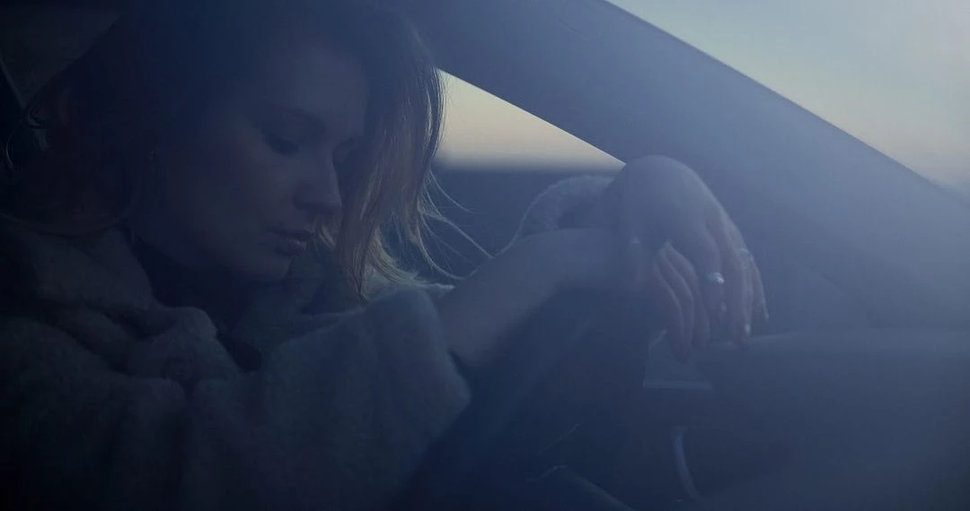 Blonde woman with her head down sitting in a car, wearing a jacket with her hands in her lap in a dimly lit setting.