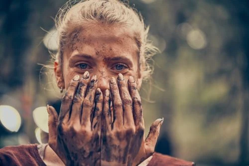 Close-up portrait of a blond woman covering her face with hands covered in mud, outdoors in a forest-like environment.