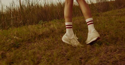 A person walking on grass in white sneakers and striped socks in a natural, outdoor environment with tall grass in the background.
